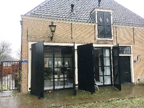 A brick building with large black shutters open to reveal expansive glass windows, featuring a dark tiled roof and an ornate upper window with diamond-patterned shutters.