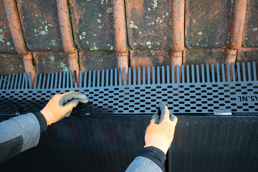 Gloved hands installing a black bird comb with perforations under mossy roof tiles.