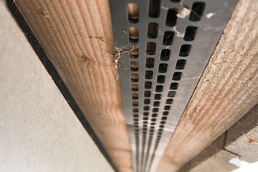Perforated metal vent between wood and a textured wall, with cobwebs.