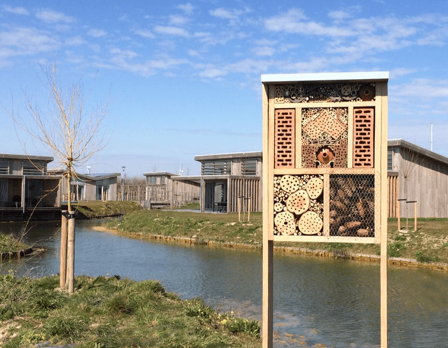A wooden insect hotel filled with various materials stands by a canal with modern wooden cabins in the background.