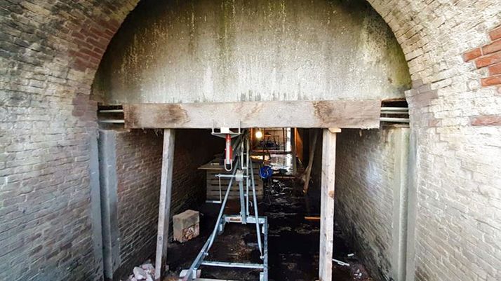 An arched brick tunnel with a large wooden beam supported by temporary wooden posts and a hydraulic jack.