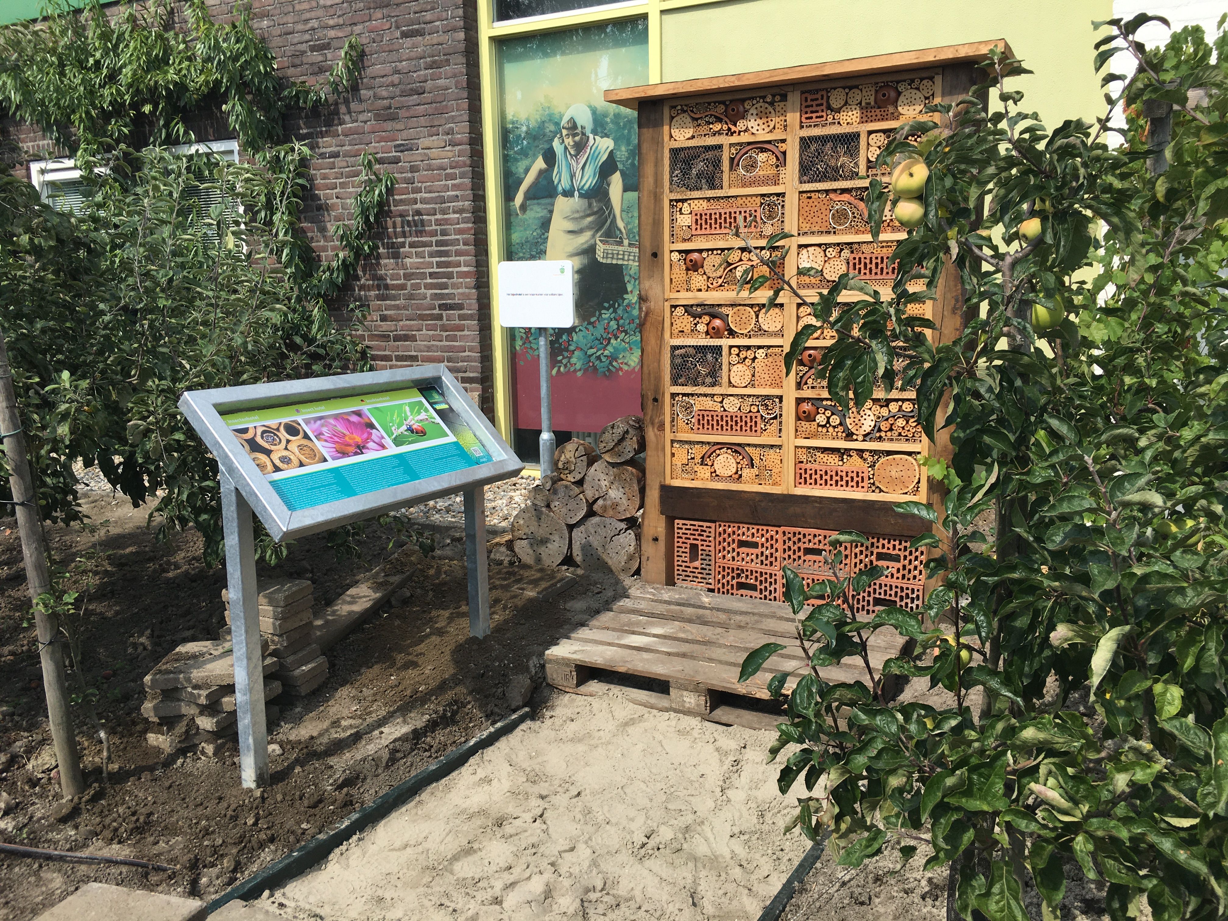 A large insect hotel and an interpretive sign stand in an outdoor garden, with an apple tree and a building in the background.