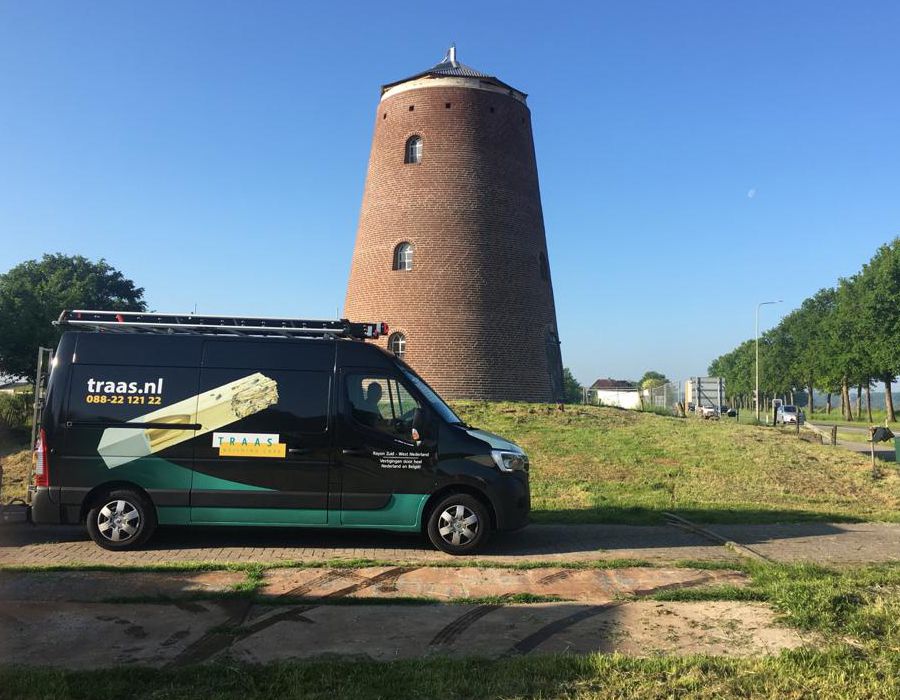 A black commercial van with "traas.nl" branding parked in front of a round brick tower on a sunny day.