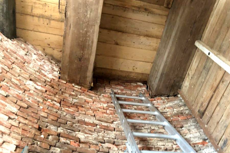 An old curved brick wall meets wooden beams and planks, with a silver ladder resting against it.