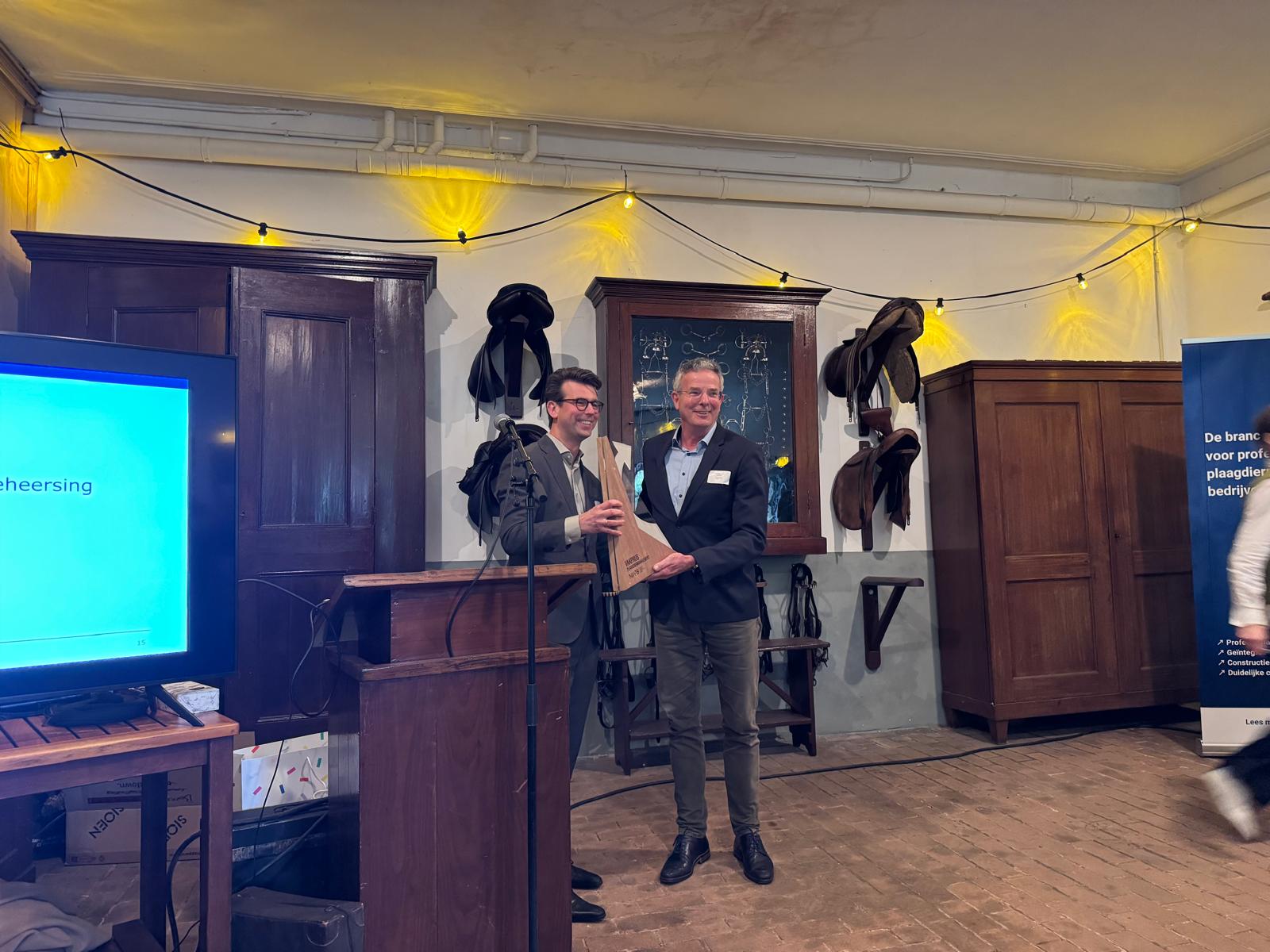 Two men on a stage, one presenting a light wooden award to the other, at an indoor event with string lights and vintage decor.