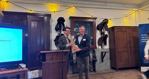 Two men on a stage, one presenting a light wooden award to the other, at an indoor event with string lights and vintage decor.