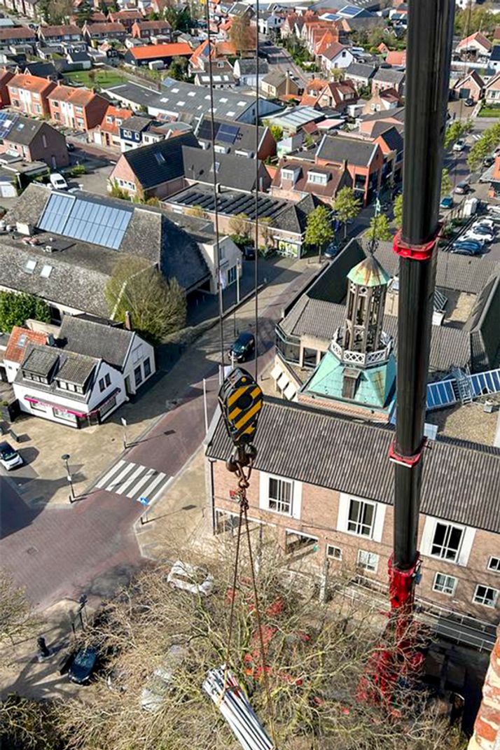 Aerial view of a crane lowering bundled pipes over bare trees in a town with houses and a church spire.