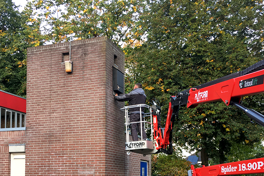 A worker in a red aerial lift installs a panel on a brick building.