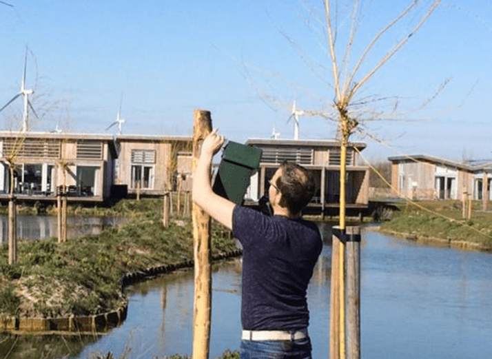 A person attaches a green box to a tree near water, with modern wooden houses and wind turbines in the background.