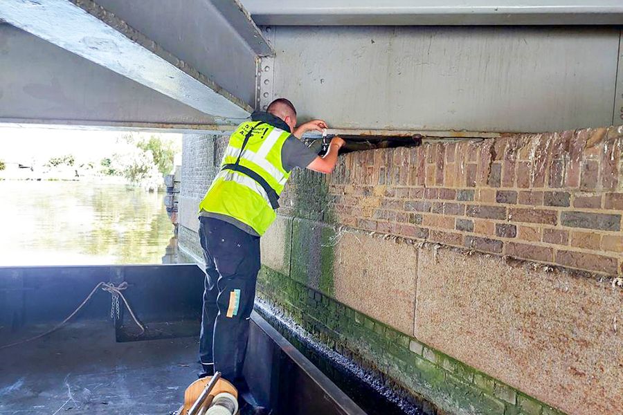 A worker in a high-vis vest uses a tool to cut into a brick wall under a bridge over water.