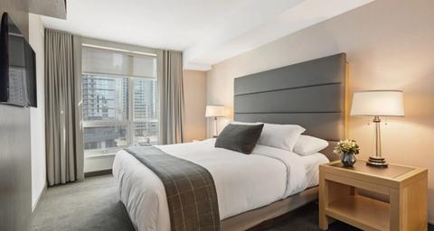 A hotel room with a bed, grey headboard, nightstand with a lamp, and a window with a city view.