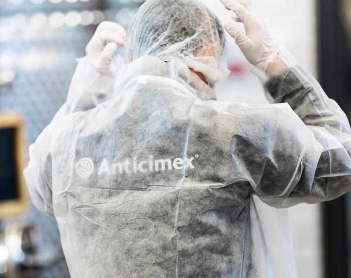 Person in white Anticimex protective suit, gloves, and hairnet adjusting headwear in an industrial facility.
