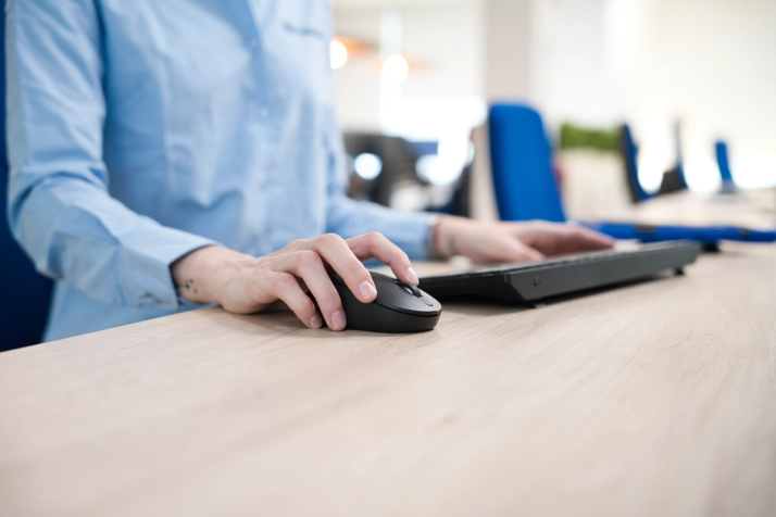 Person in a blue shirt using a computer mouse and keyboard on a wooden desk.