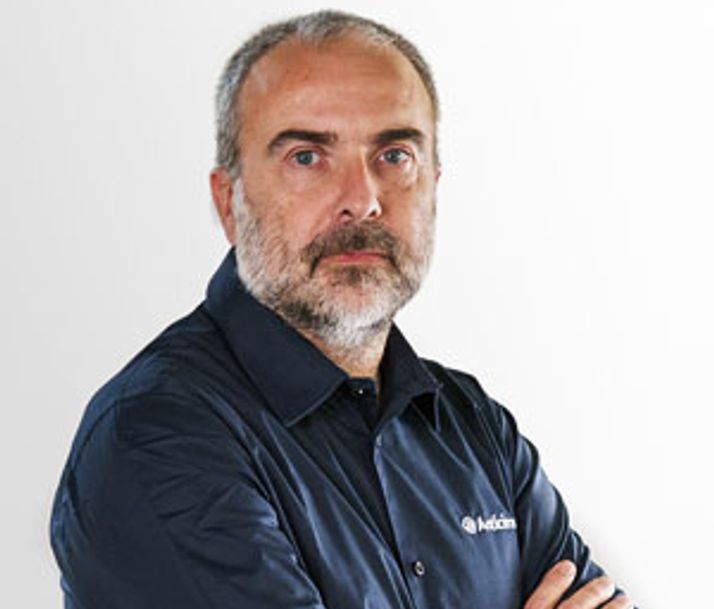 A man with a grey beard and hair, wearing a dark blue shirt with an "Avidcom" logo, crosses his arms and looks at the camera.