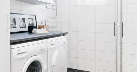 Ljust helkaklat badrum med tvättmaskin, torktumlare och duschhörna i glas.