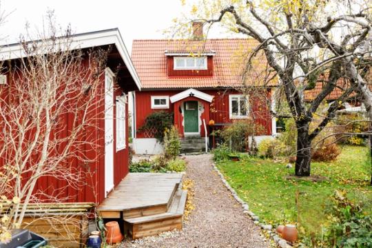 En grusad gång som leder fram till ett rött hus med vita knutar, ett attefallshus står till vänster i bild.