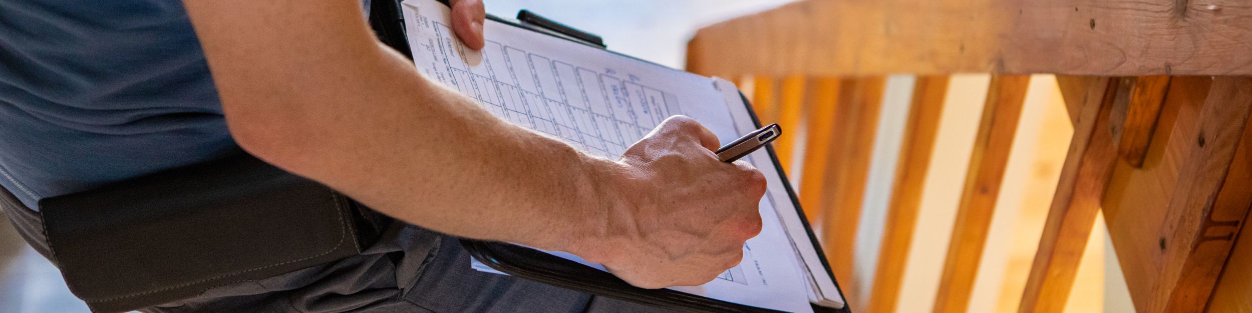 En man med mörk t-shirt och mörka byxor står vid ett trappräcke i furu med en skrivplatta och ett protokoll som fylls i