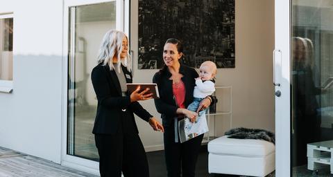 Mäklare pratar med kvinna som håller i en bebis framför vitt hus