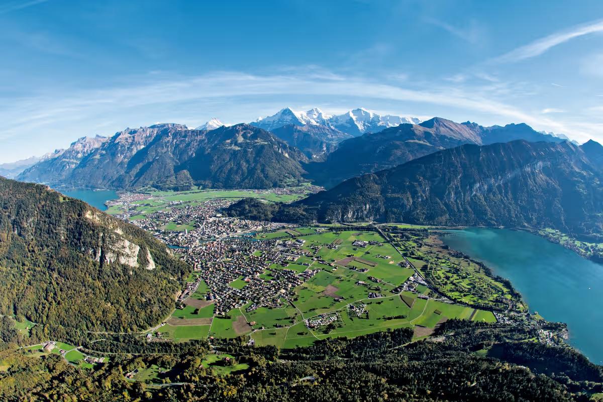 Panoramic view of a town in a valley between two lakes, surrounded by mountains with distant snow-capped peaks.