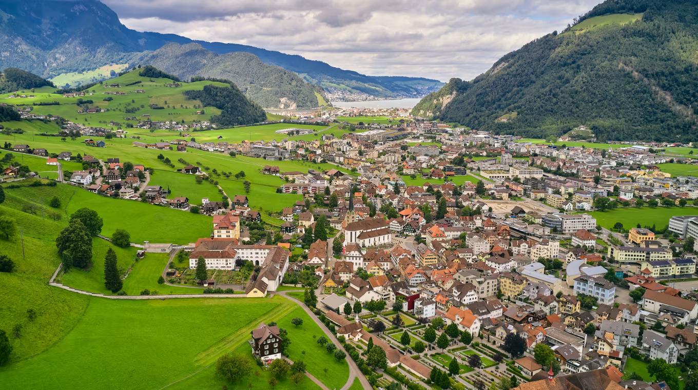 Aerial view of a town nestled in a lush green valley, surrounded by mountains, with a lake in the distance under a cloudy sky.
