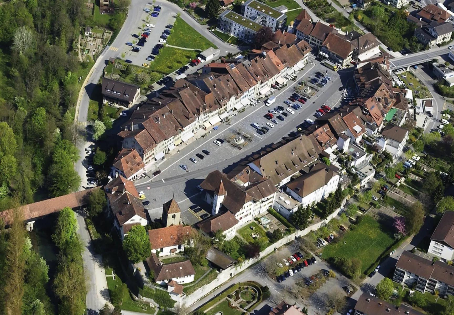 Aerial view of a historic European town with a long central square, red-roofed buildings, and a long wooden bridge extending over a green valley.