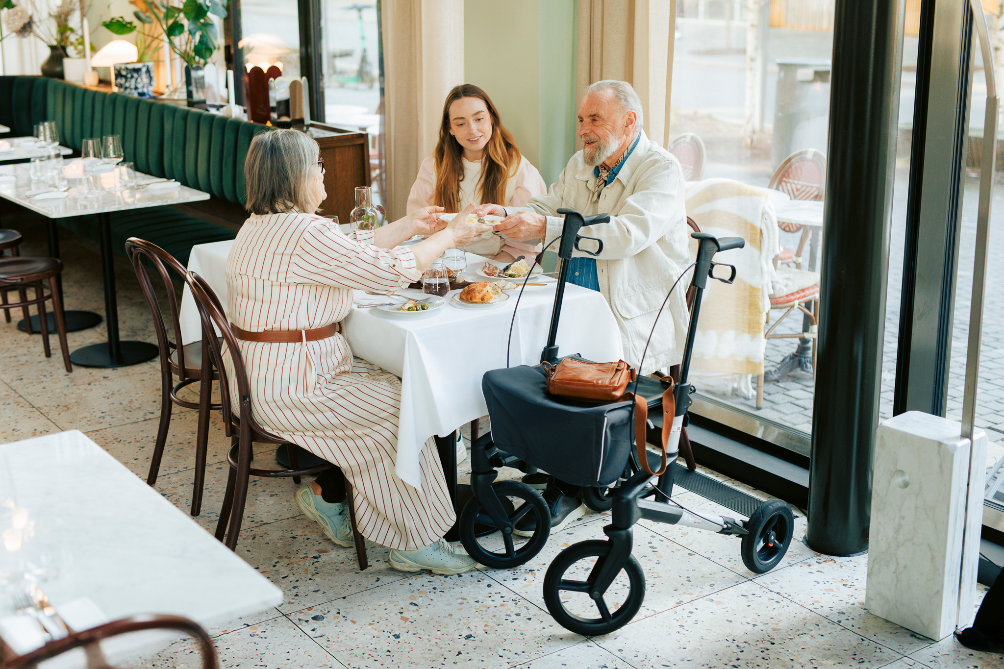 Et eldre par og en ung kvinne spiser sammen på en reataurant. Ved siden av bordet står en rullator
