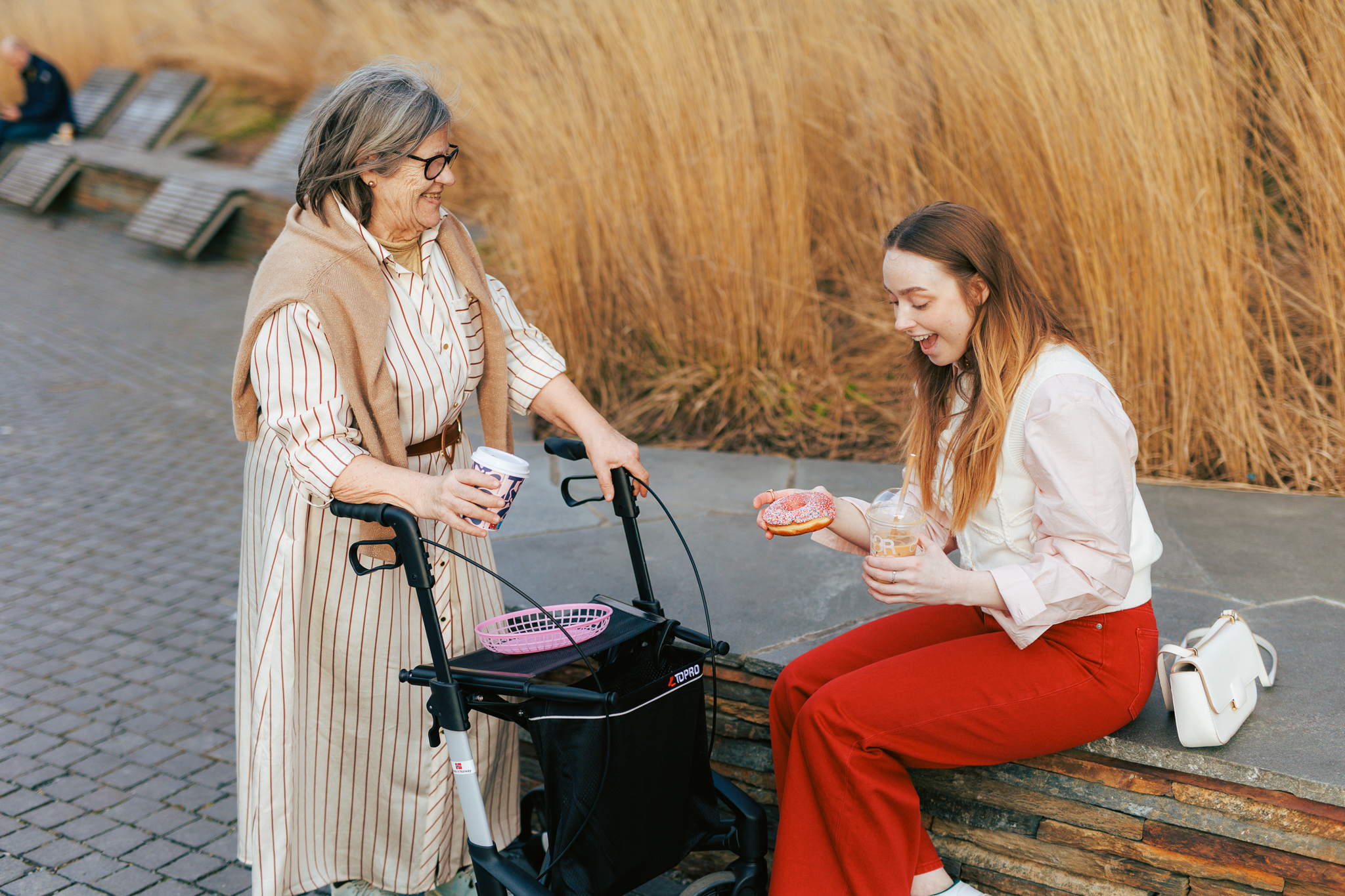 en eldre dame med rullator og en ung kvinne spiser en donut på en benk