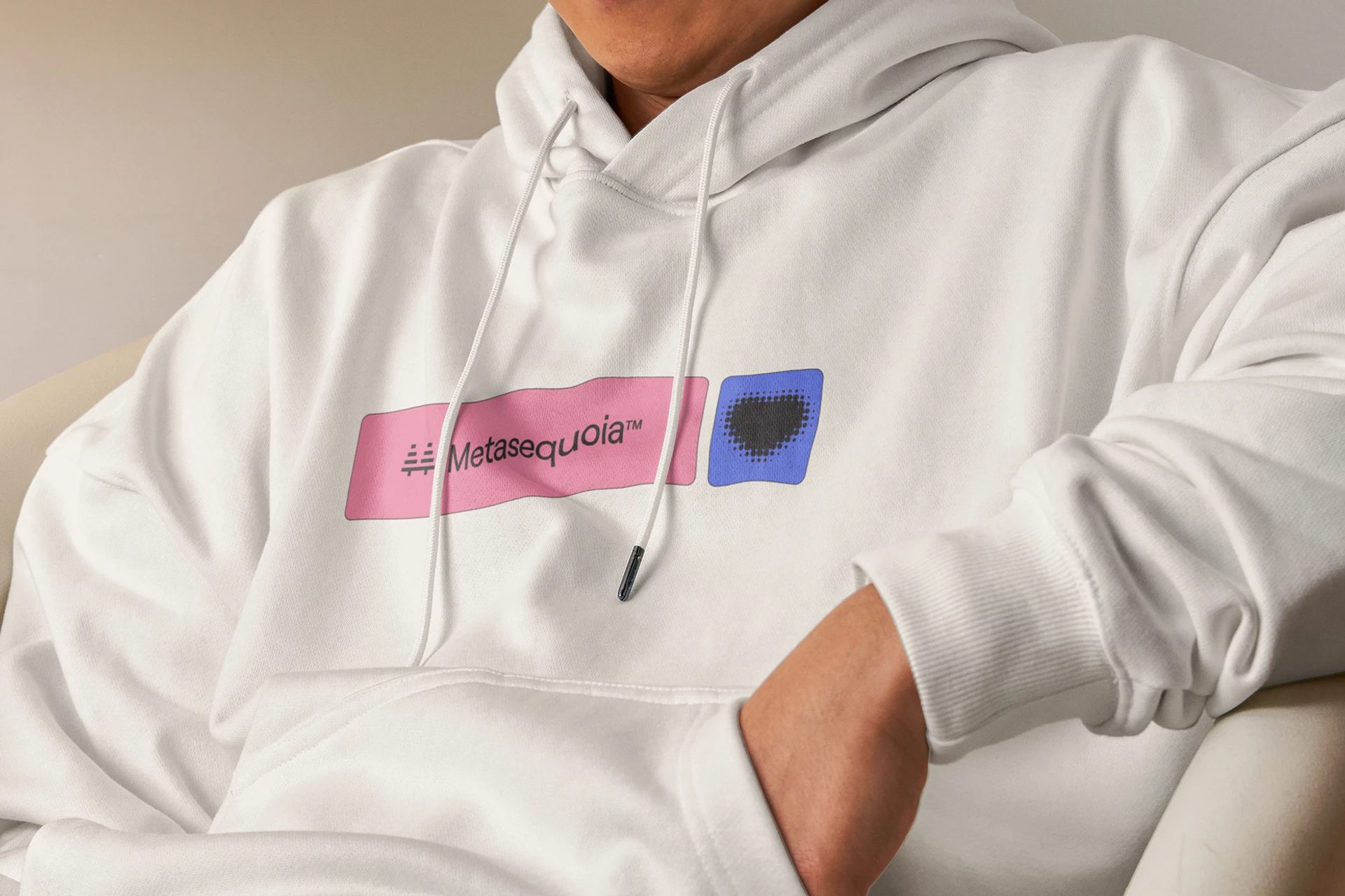 White hoodie with a pink Metasequoia™ chest graphic and a blue square, worn by a seated person against a beige background.