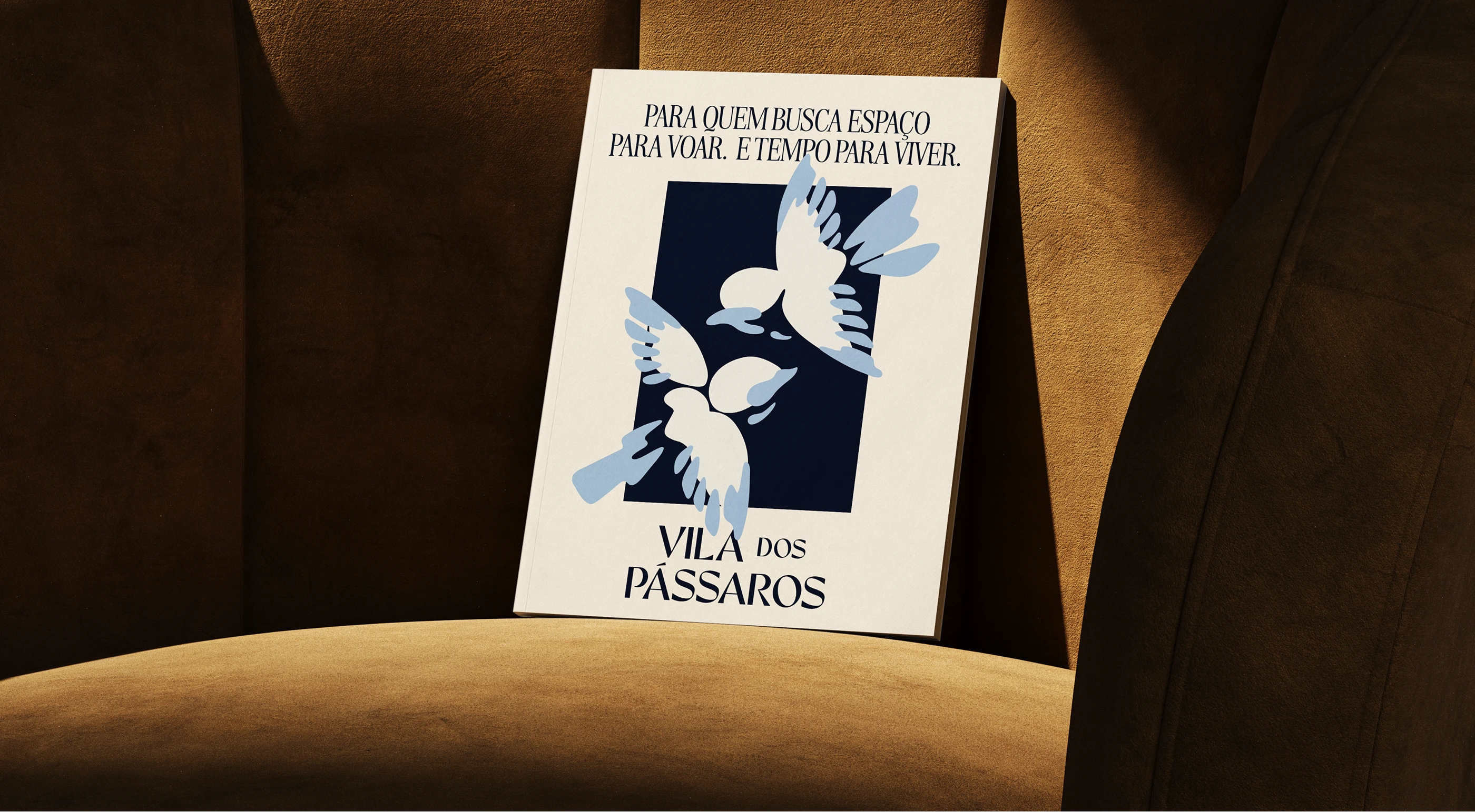 Cream Vila dos Pássaros brochure with blue birds and Portuguese text, propped on a sunlit brown chair.