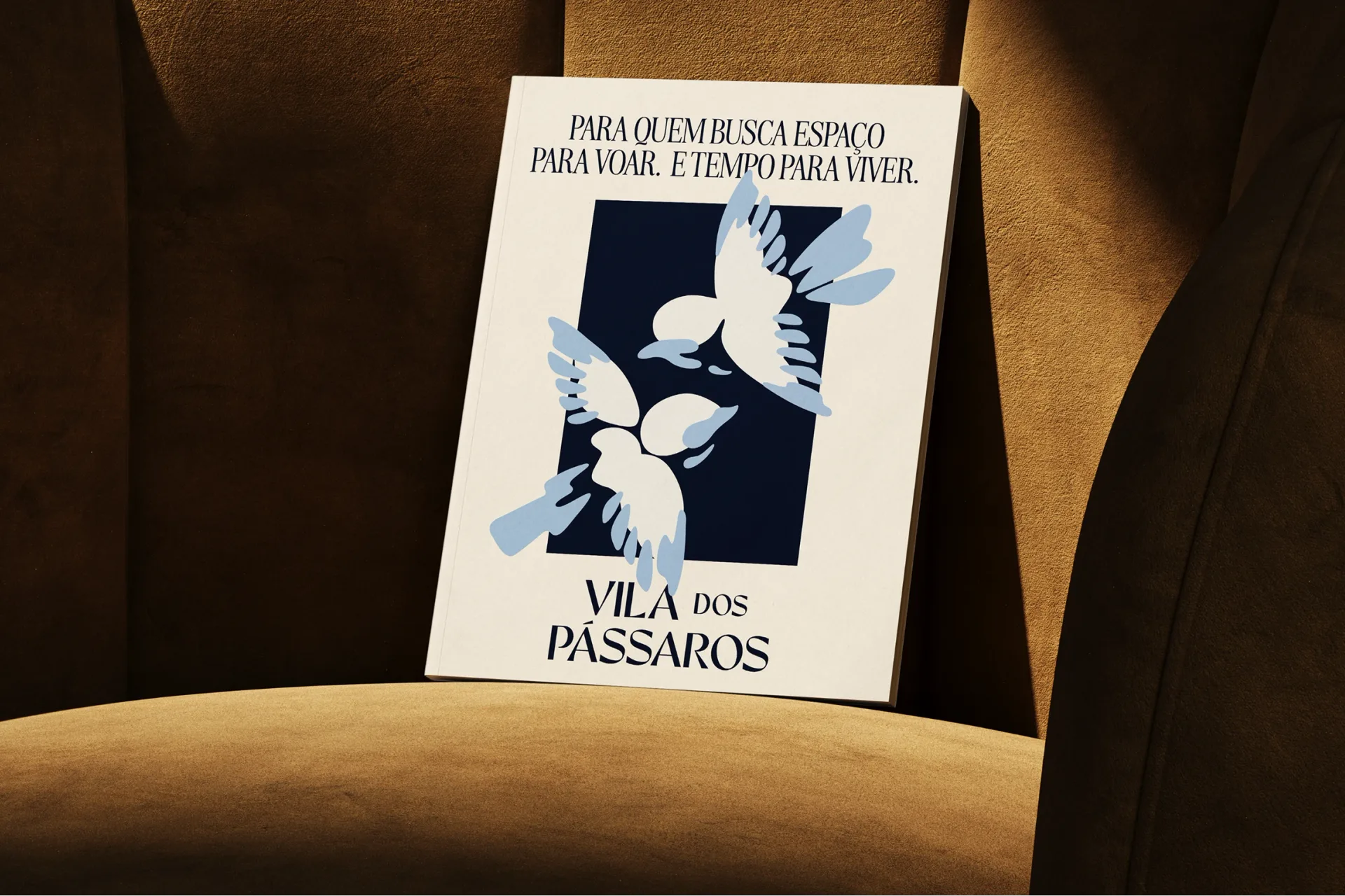 Cream Vila dos Pássaros brochure with blue birds and Portuguese text, propped on a sunlit brown chair.