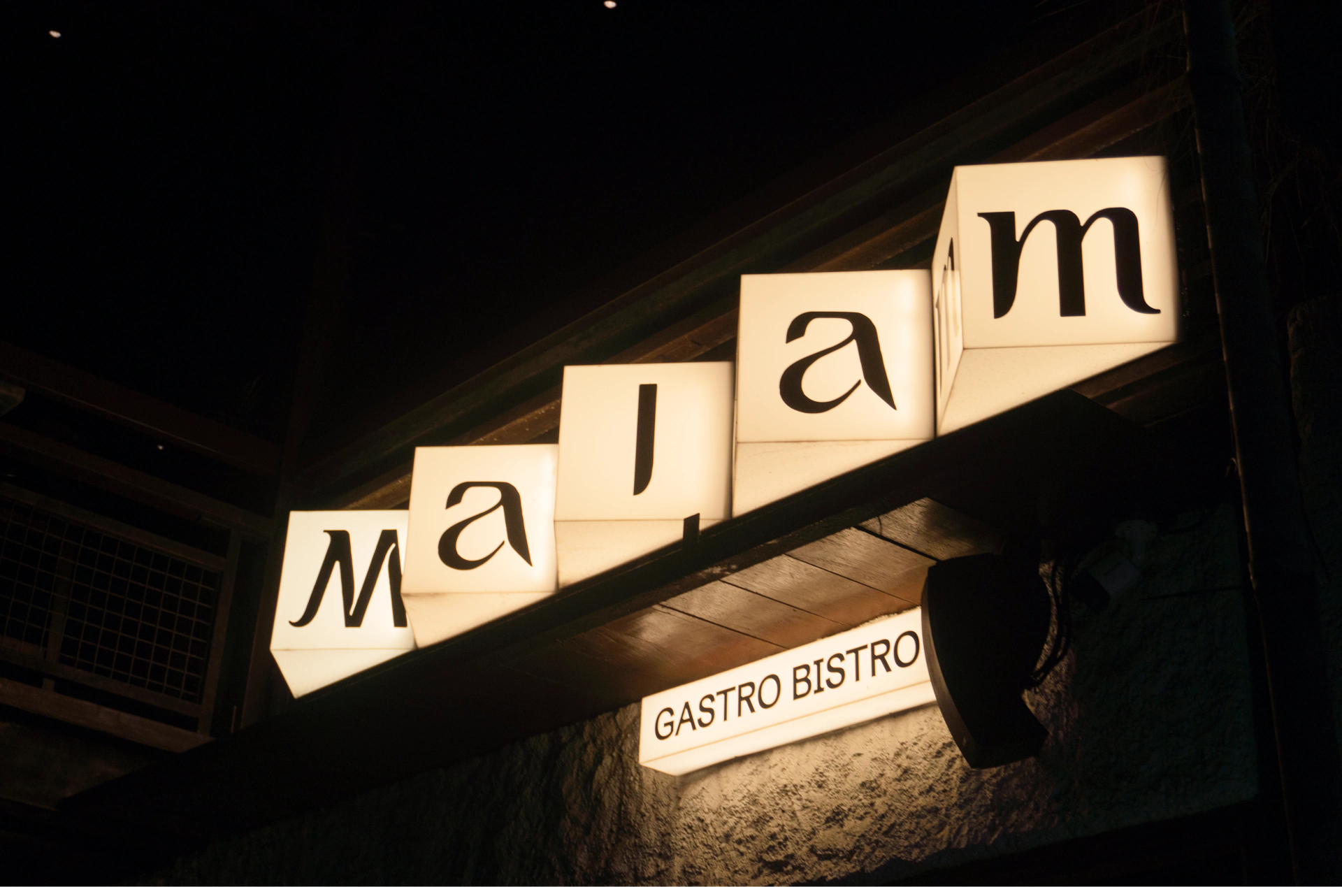 Illuminated cream cube signage spells “Malam” in black above a rectangular “GASTRO BISTRO” sign on a dark facade.