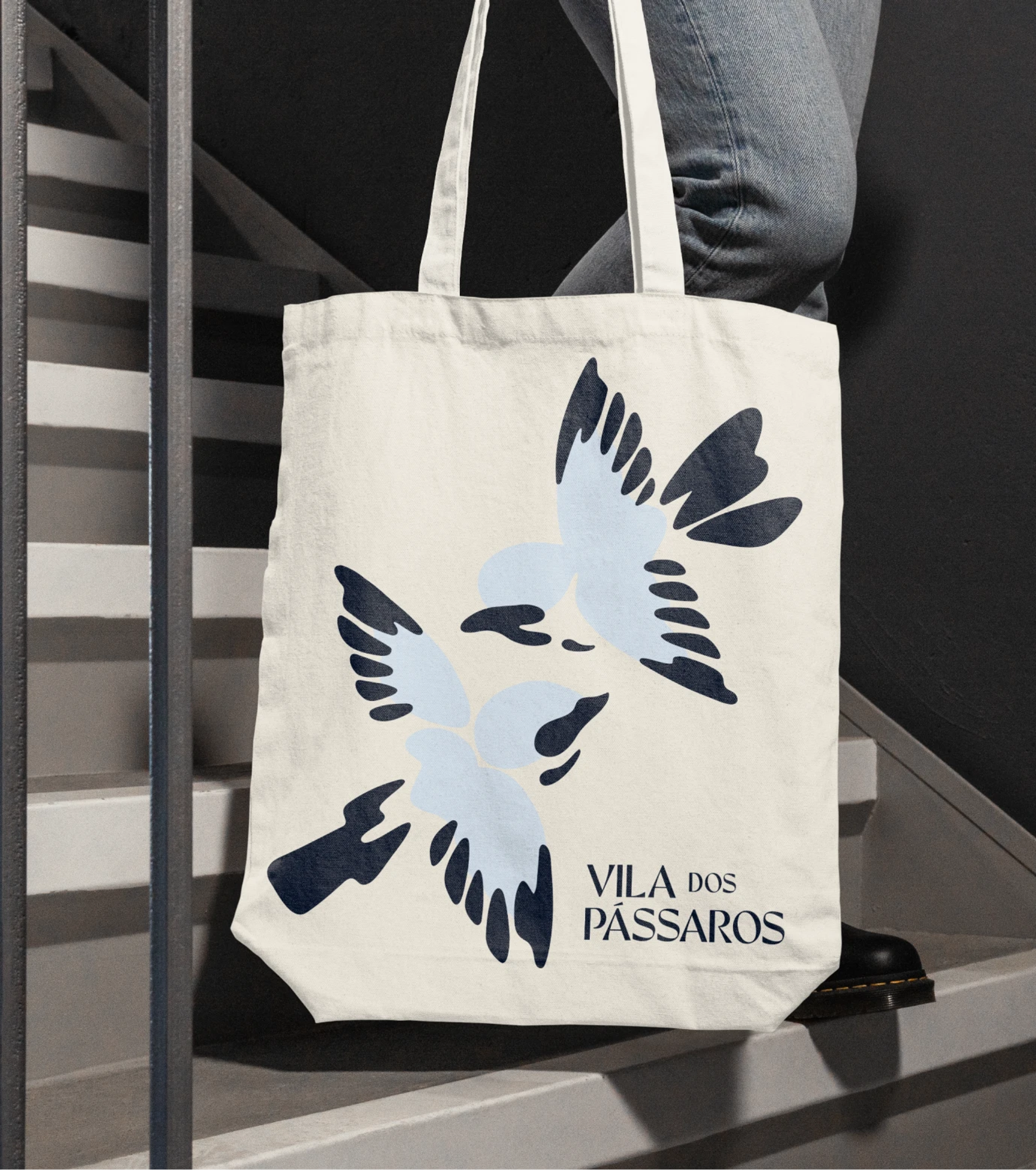 Cream tote bag with pale blue and navy birds and “VILA DOS PÁSSAROS” text, carried by a person on grey stairs.