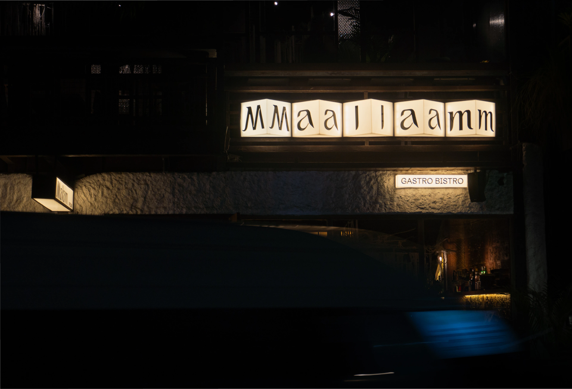 Illuminated cream signage reading “Malam” and “GASTRO BISTRO” spans a dark stone restaurant facade at night.