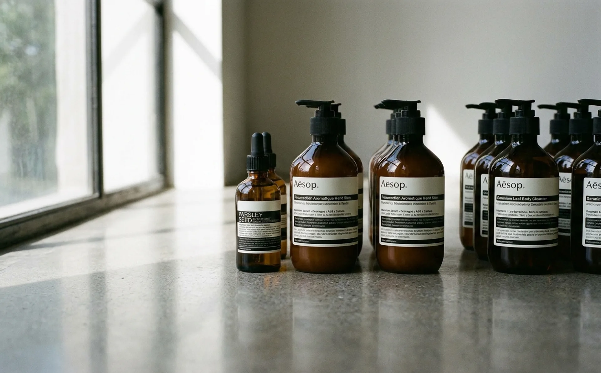 Aesop amber apothecary bottles with minimal black-and-white labels arranged on a marble surface, lit by soft natural side light