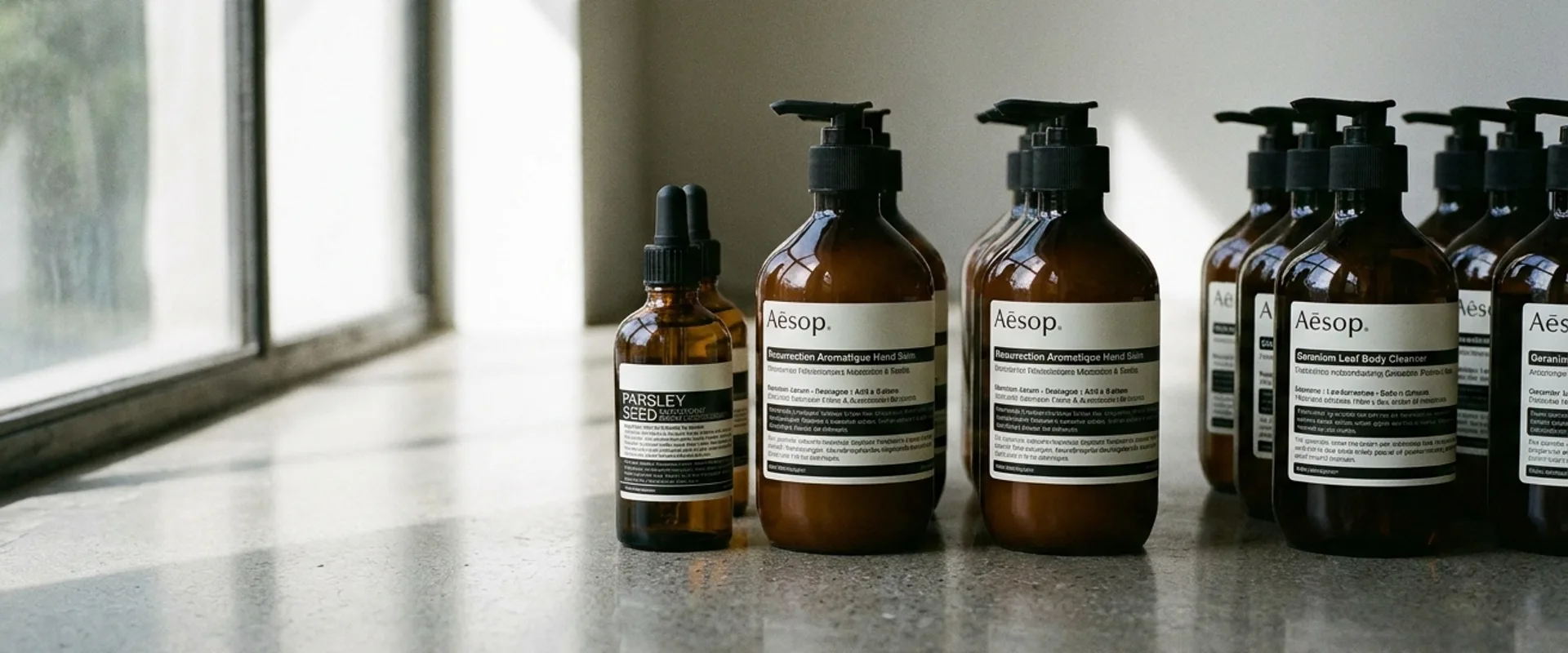 Aesop amber apothecary bottles with minimal black-and-white labels arranged on a marble surface, lit by soft natural side light