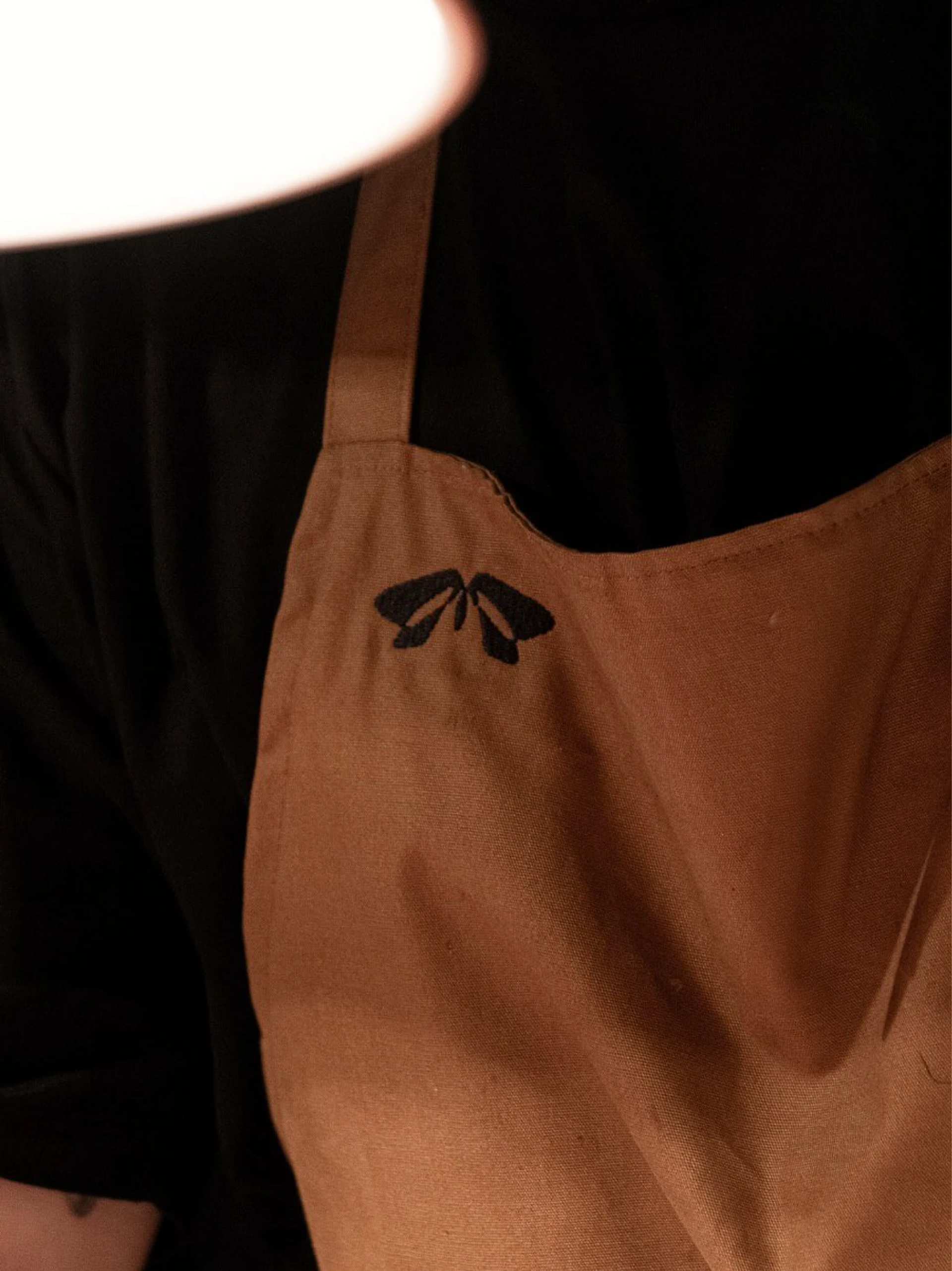 Close-up of a brown apron with a black embroidered moth emblem, worn over a black shirt beneath a white lampshade.