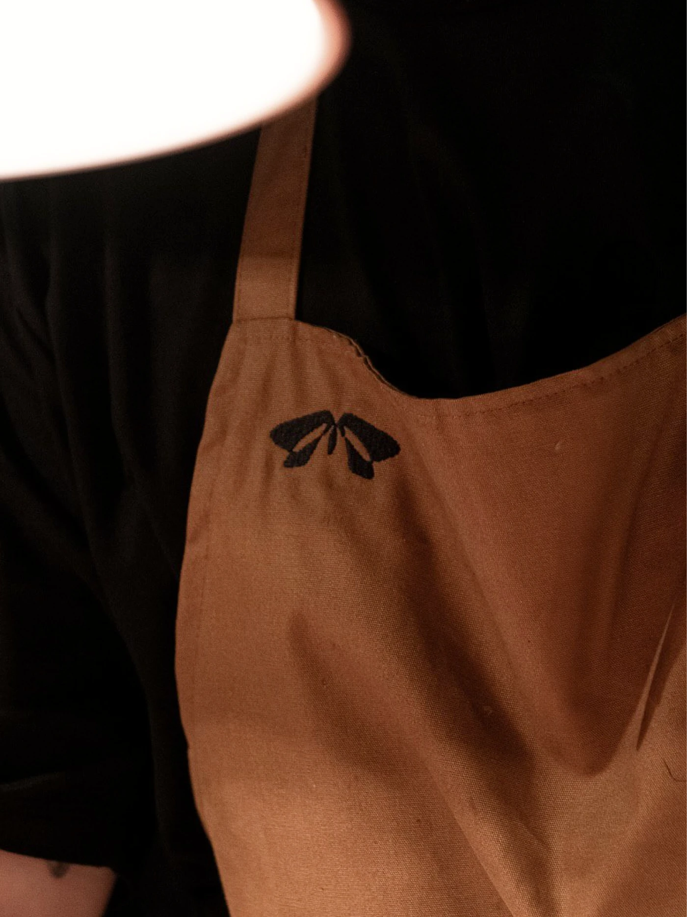 Close-up of a brown apron with a black embroidered moth emblem, worn over a black shirt beneath a white lampshade.