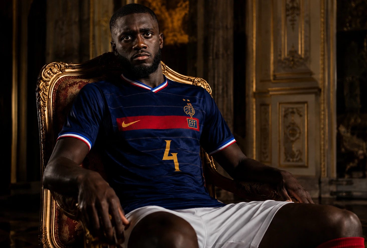 Footballer in a navy France jersey marked F.F.F and 4 sits on a gold chair in an ornate gold and brown interior.
