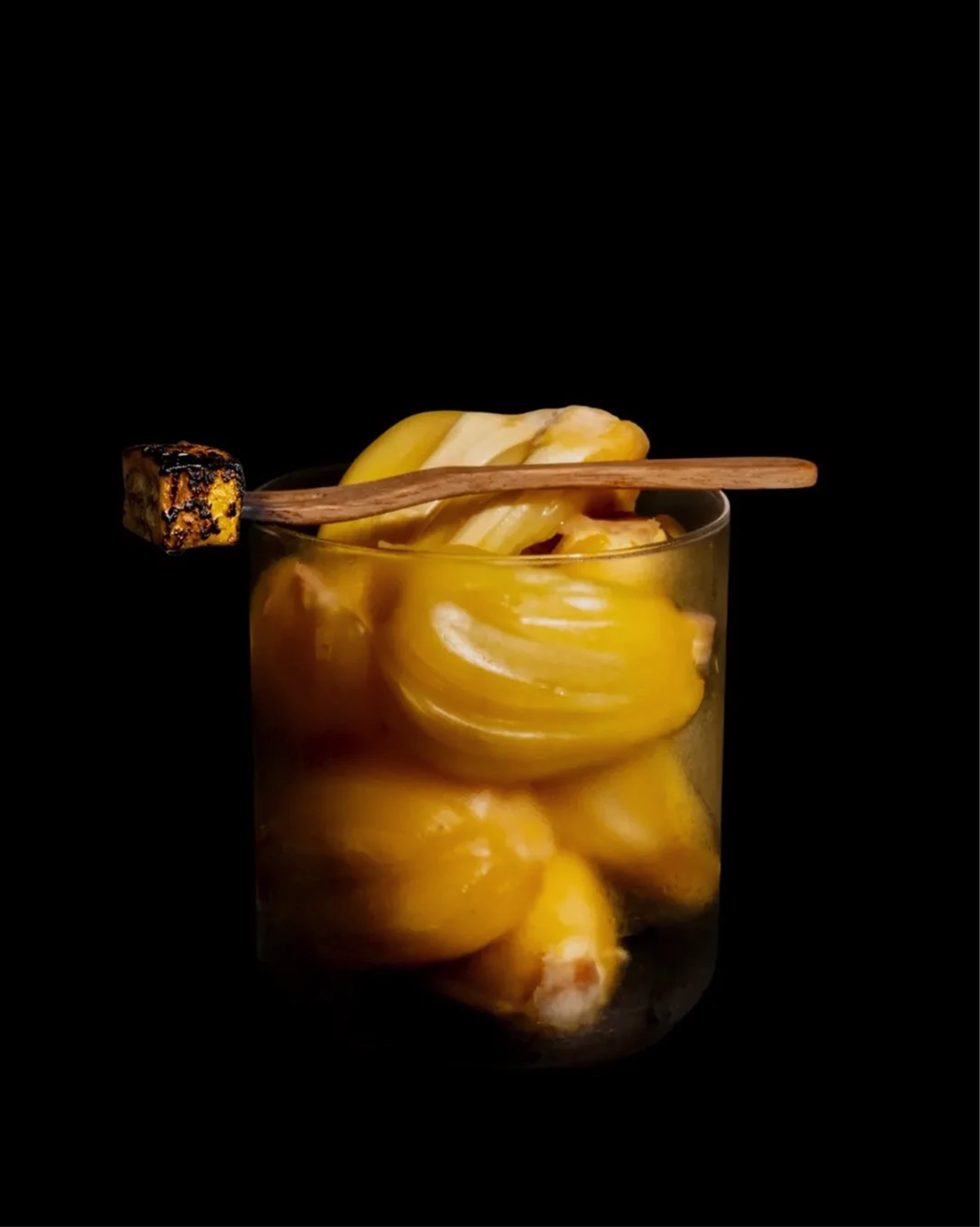 Clear glass filled with yellow glazed pastries, topped by a wooden skewer with a charred cube, against black.