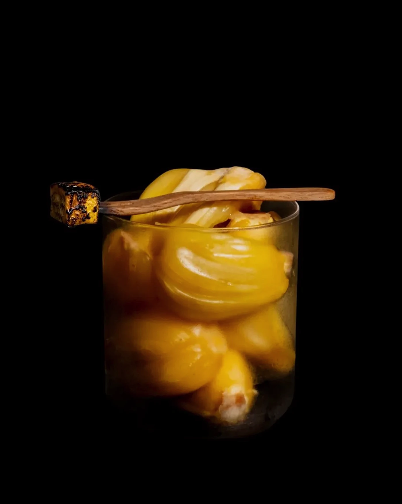 Clear glass filled with yellow glazed pastries, topped by a wooden skewer with a charred cube, against black.
