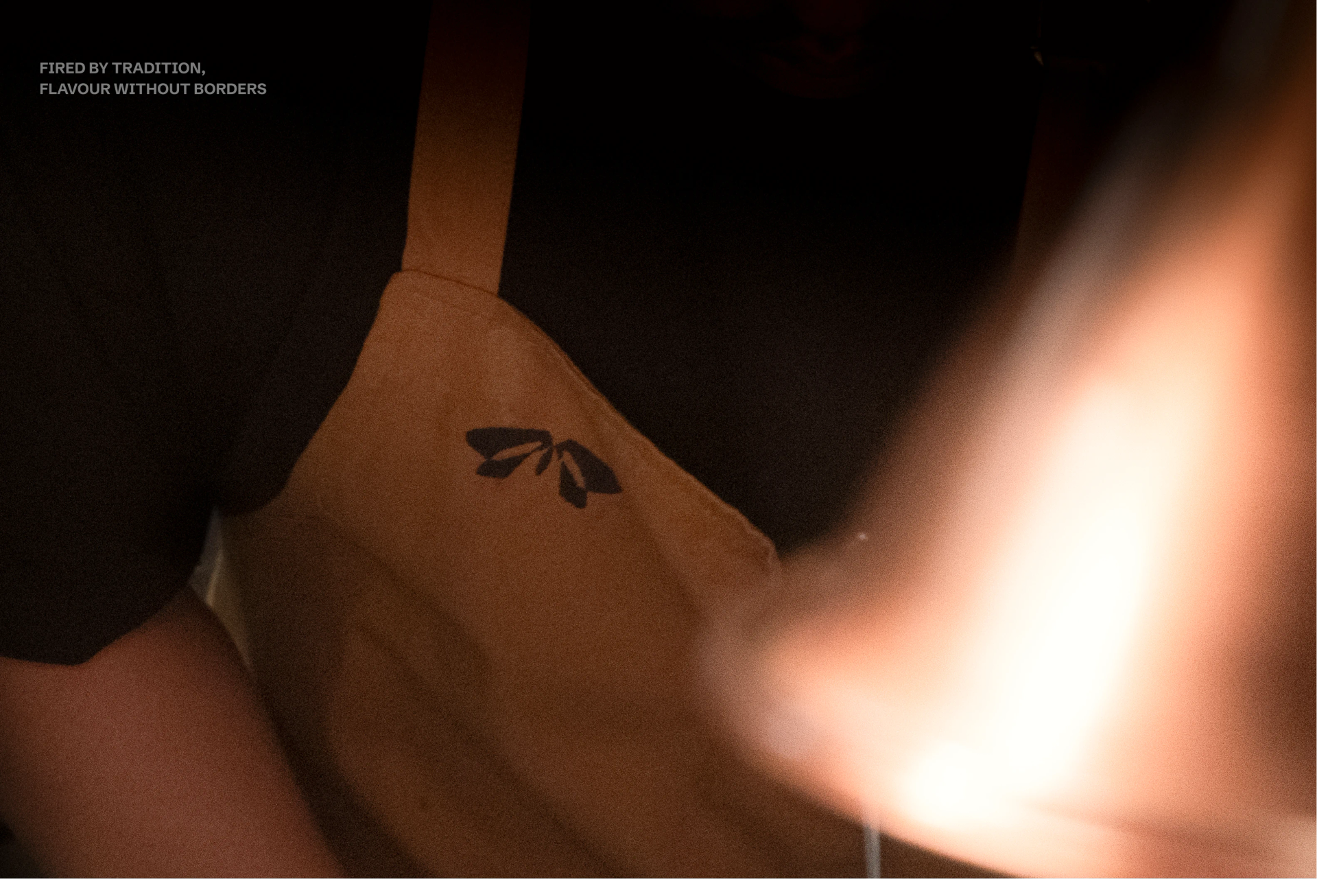 Brown apron with a black leaf mark beside orange flames, with “FIRED BY TRADITION, FLAVOUR WITHOUT BORDERS” in white.