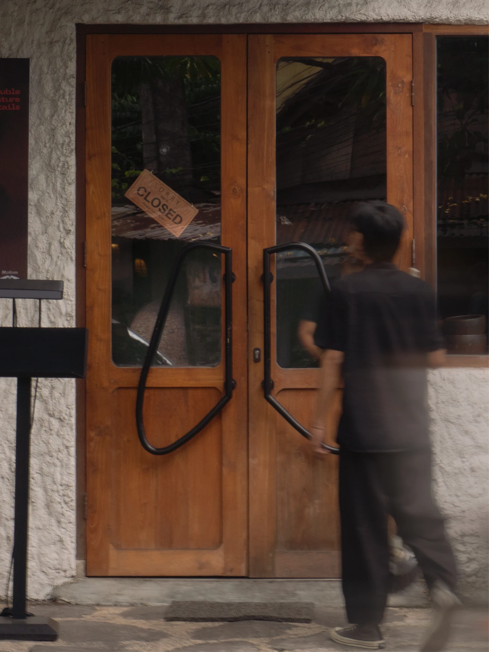 Wooden bistro doors with black loop handles and a brown “SORRY CLOSED” sign, as a blurred person passes in front.