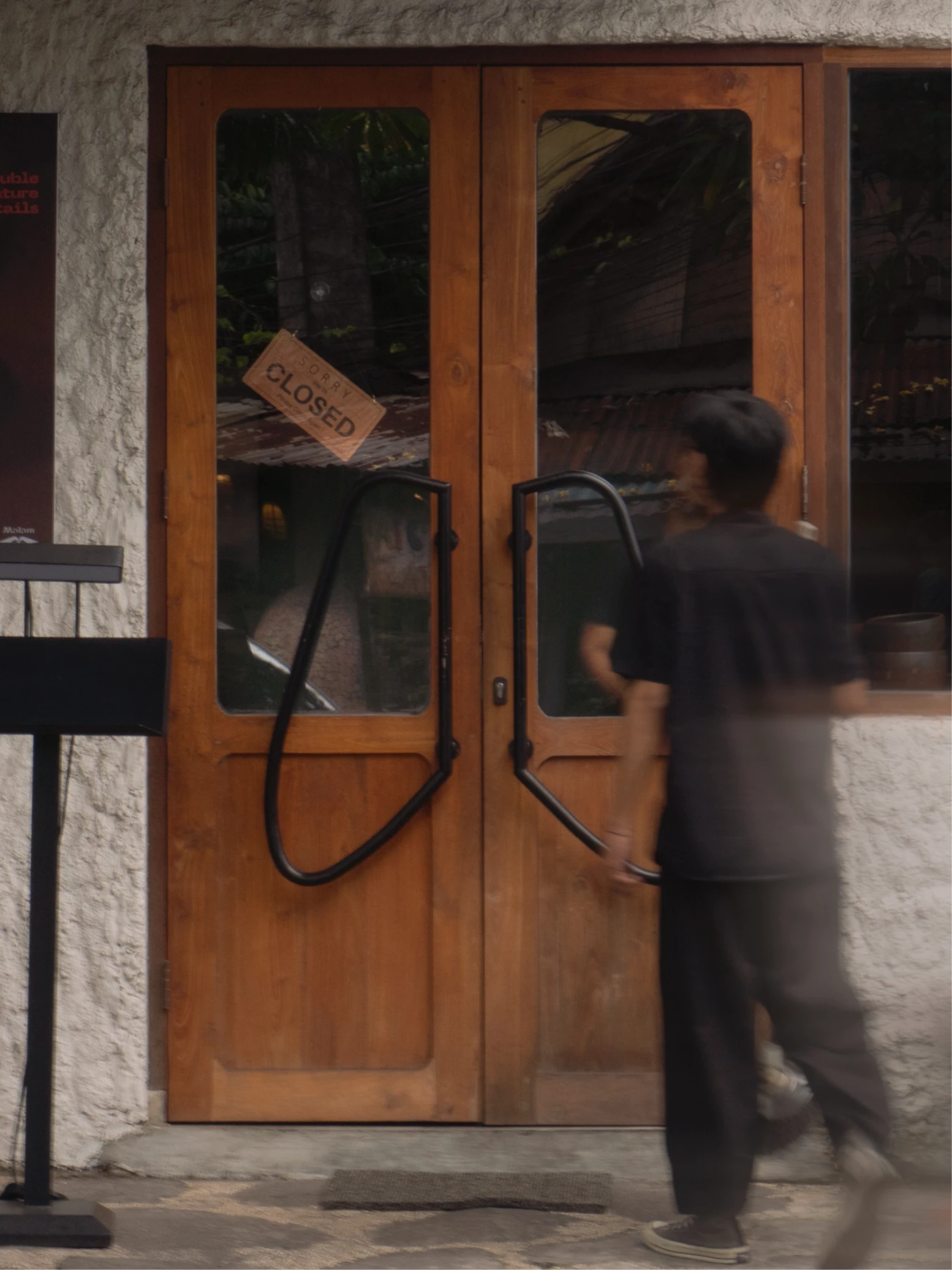 Wooden bistro doors with black loop handles and a brown “SORRY CLOSED” sign, as a blurred person passes in front.