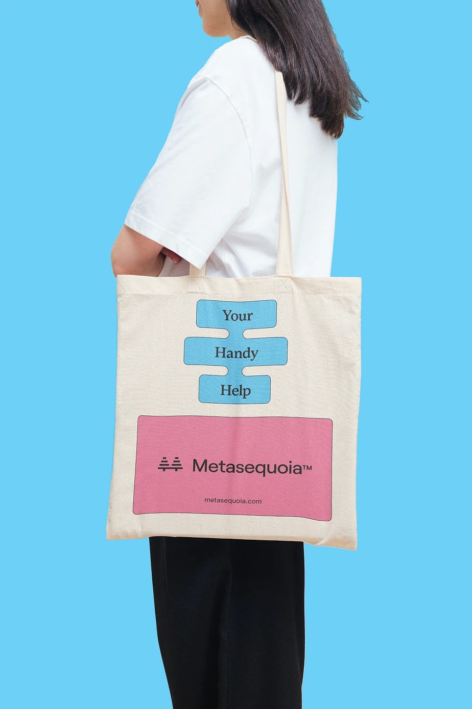 Cream tote bag with blue “Your Handy Help” blocks and a pink “Metasequoia™” panel, carried against a blue background.