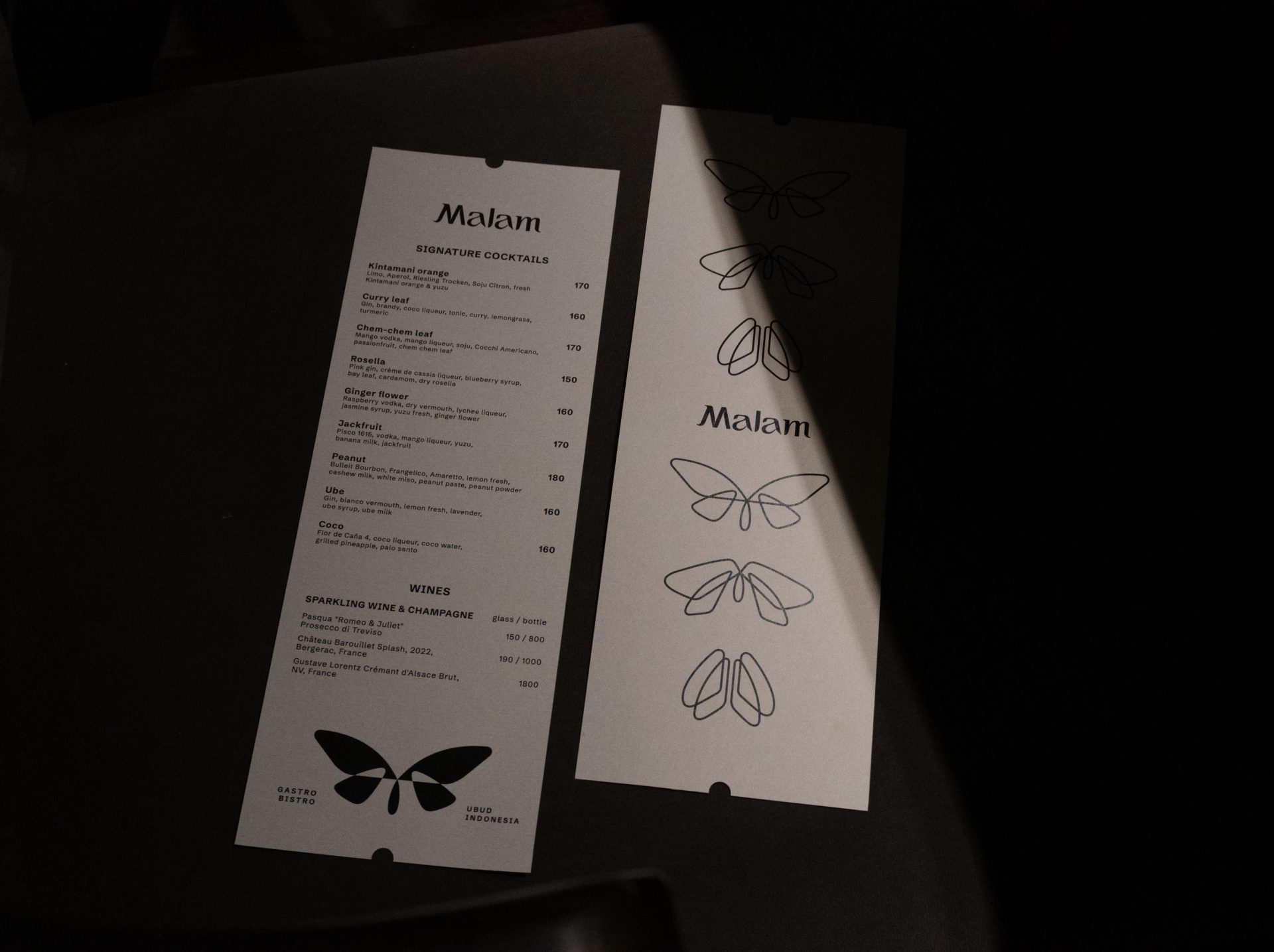 Two white menu cards show “Malam” cocktail and wine listings with black butterfly graphics, arranged on a dark surface.