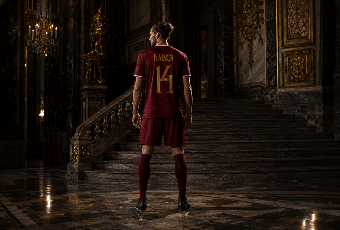 Footballer in a burgundy kit marked “RABIOT 14” in gold stands before a marble staircase in an ornate dark interior.