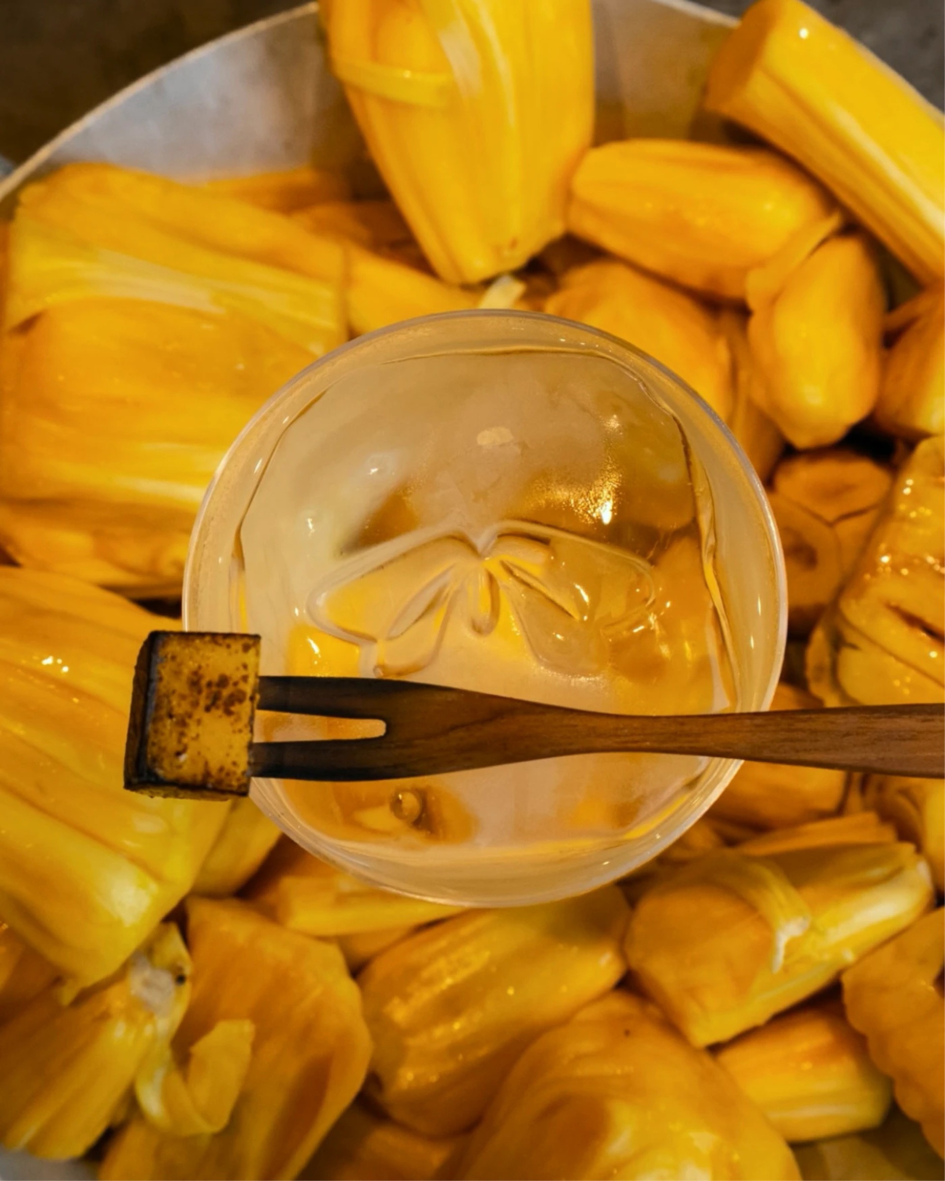 Top-down view of a clear iced drink and wooden cocktail pick arranged over a tray of yellow jackfruit pods.