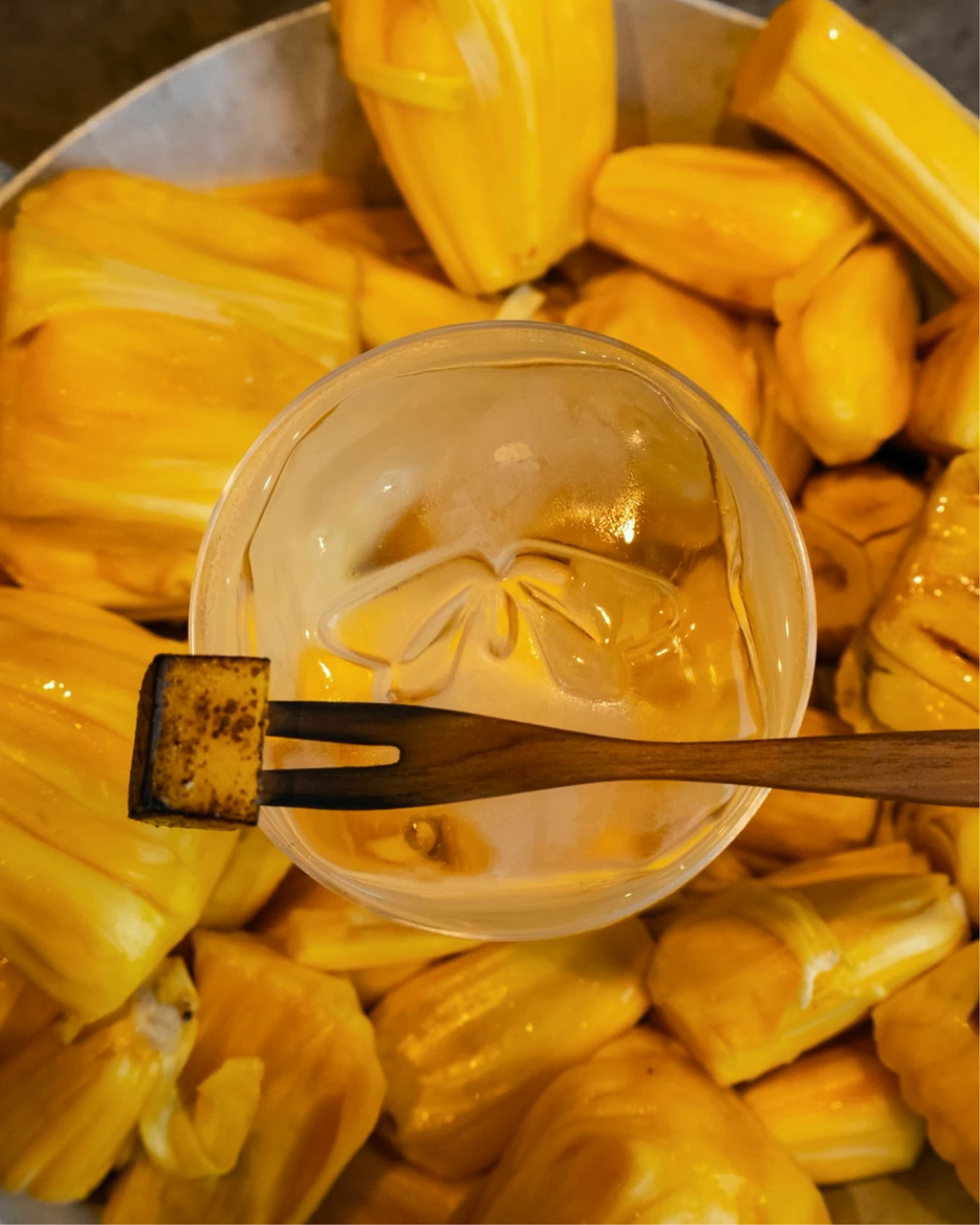 Top-down view of a clear iced drink and wooden cocktail pick arranged over a tray of yellow jackfruit pods.