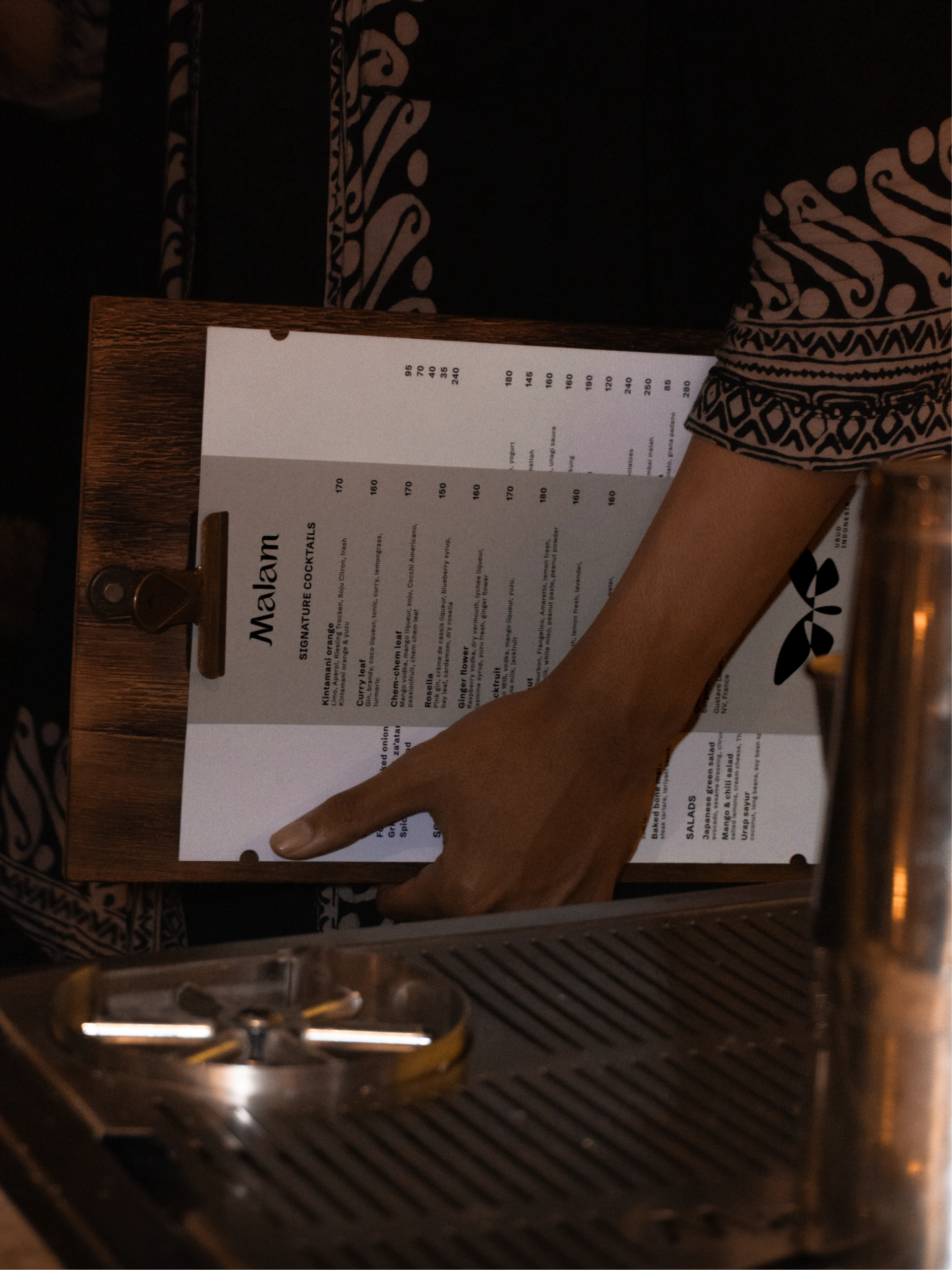 A hand holds a wooden clipboard with a black-and-white Malam SIGNATURE COCKTAILS menu beside metal bar equipment.