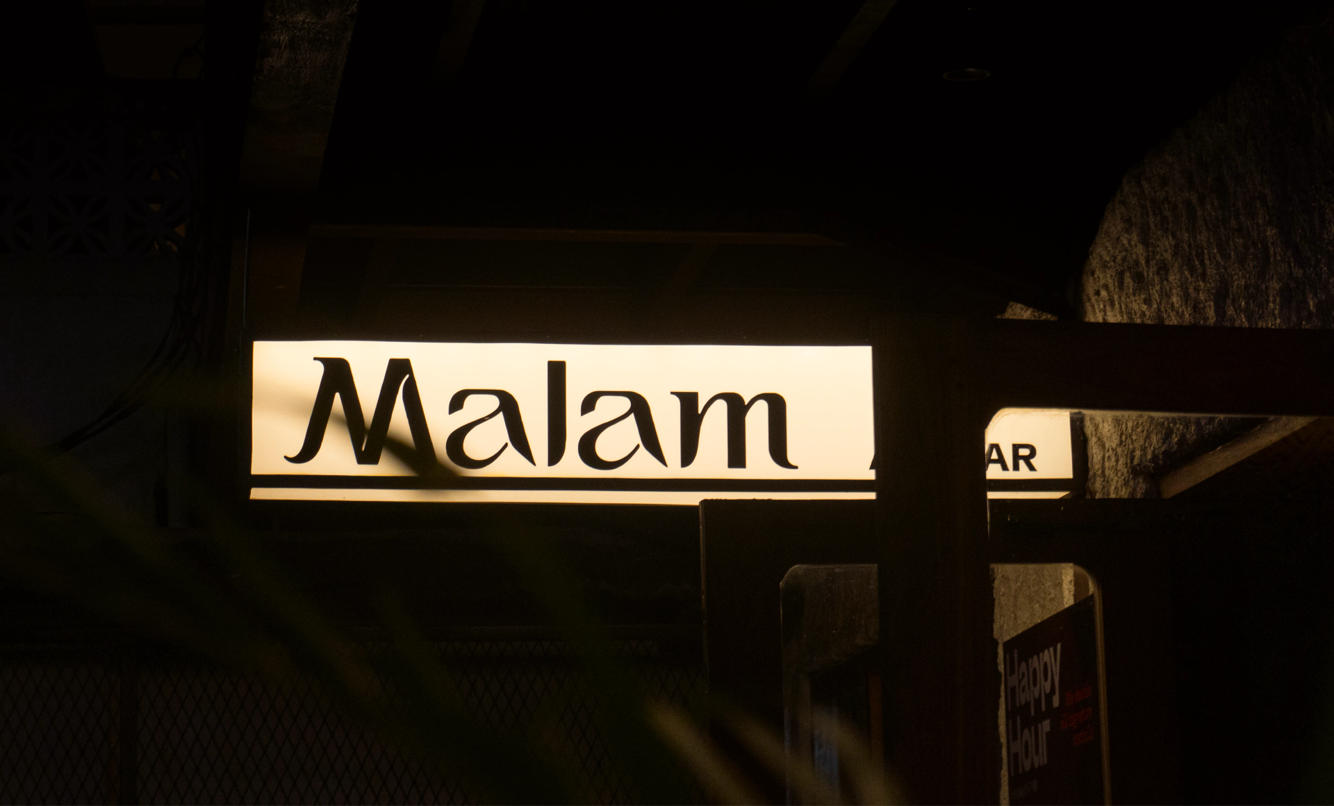 Illuminated cream storefront signage reads “Malam” in black lettering, framed by a dark entrance and stone walls at night.